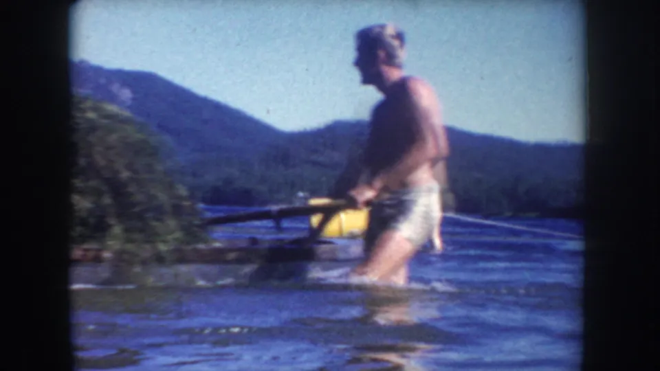 Adult man in swim trunks wades through a South Dakota lake in 1954, with a yellow buoy, wooded shore, and hills behind him.