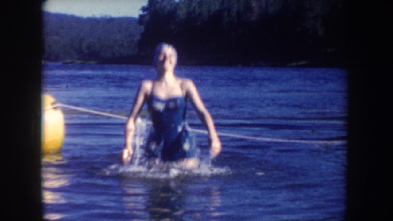 Adult woman in a dark one-piece swimsuit and swim cap moves through South Dakota lake water, with buoy hardware and shoreline behind her.