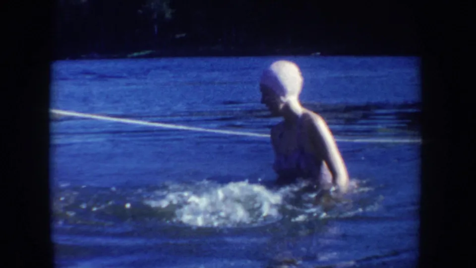 1954 South Dakota home-movie frame showing a woman in a light swim cap and dark swimsuit standing after a splash in the roped lake area.