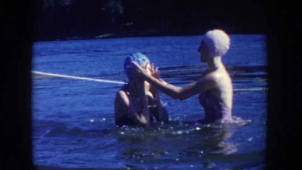1954 South Dakota lake frame where a woman in a light cap helps adjust the blue cap of another swimmer in the water.