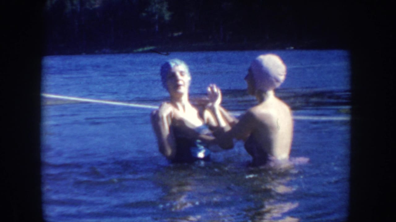 1954 South Dakota home-movie frame of two women in swim caps and swimsuits standing in lake water, one helping adjust the other's cap.