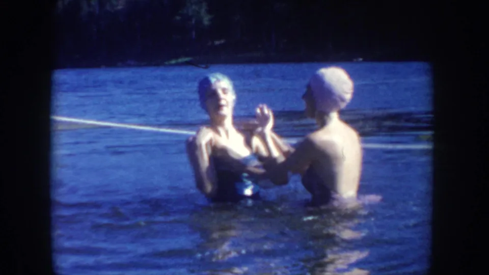 1954 South Dakota home-movie frame of two women in swim caps and swimsuits standing in lake water, one helping adjust the other's cap.