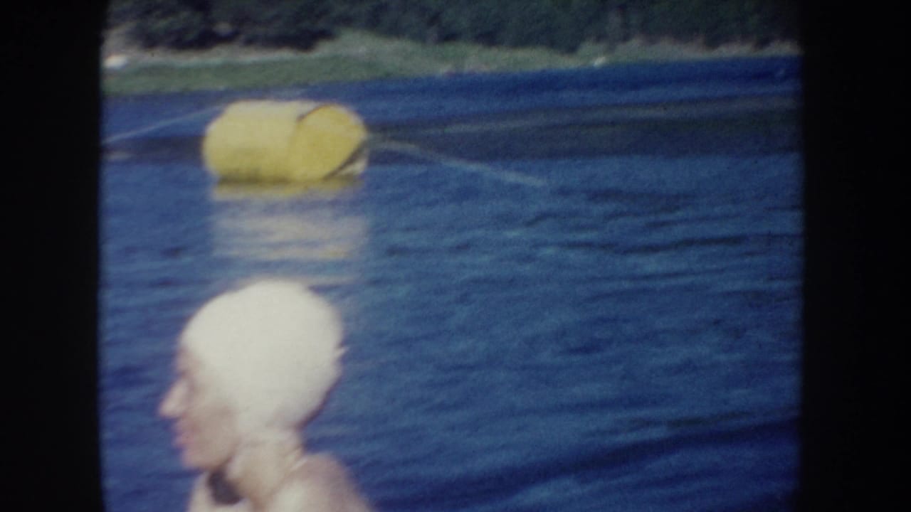 1954 South Dakota home-movie frame with a yellow buoy floating on the lake and a swim-capped woman partly visible in the foreground.