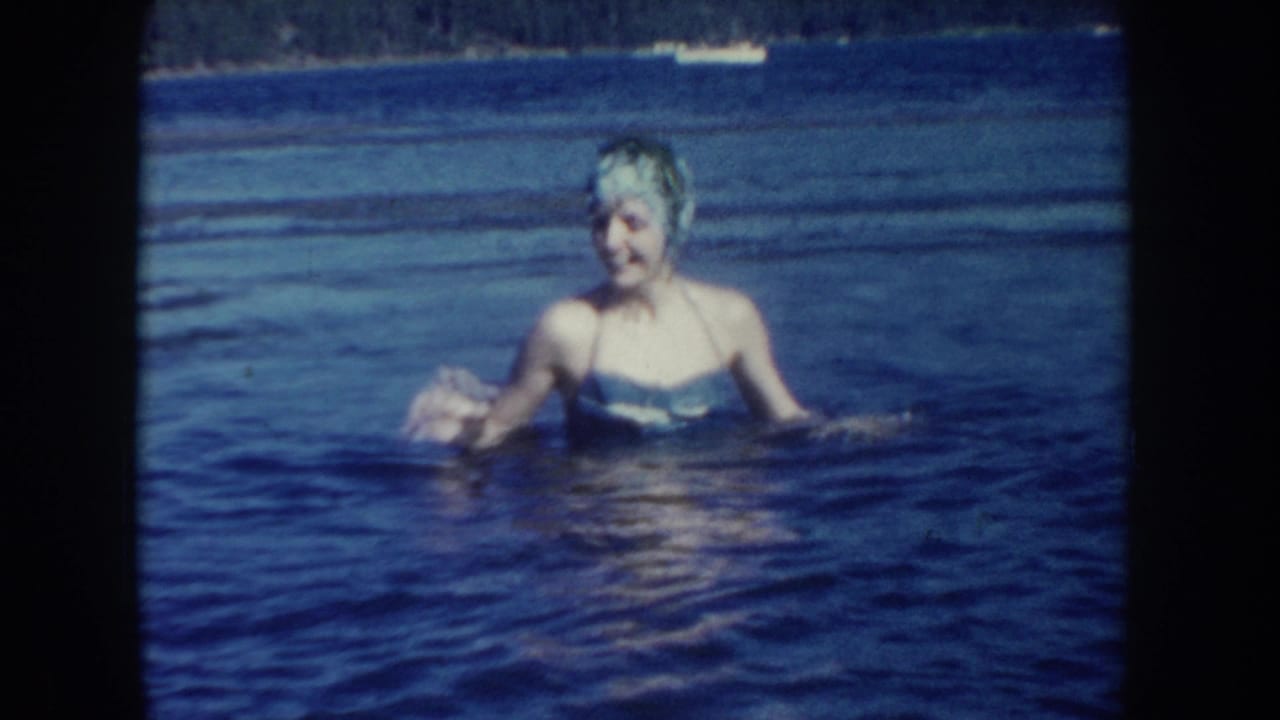 1954 South Dakota frame of a woman in a swim cap and one-piece swimsuit wading in a lake, with shoreline and small boats behind her.