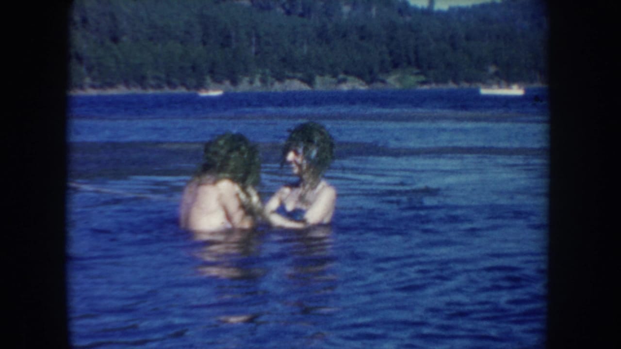 Two young women in 1950s lake footage face each other in shallow water, both partly covered with wet aquatic plants.