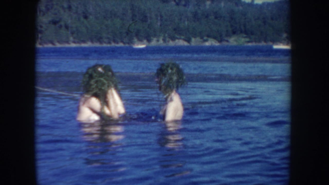 The two swimmers stand in blue lake water near a wooded shoreline, one adjusting plants or wet hair after the playful exchange.