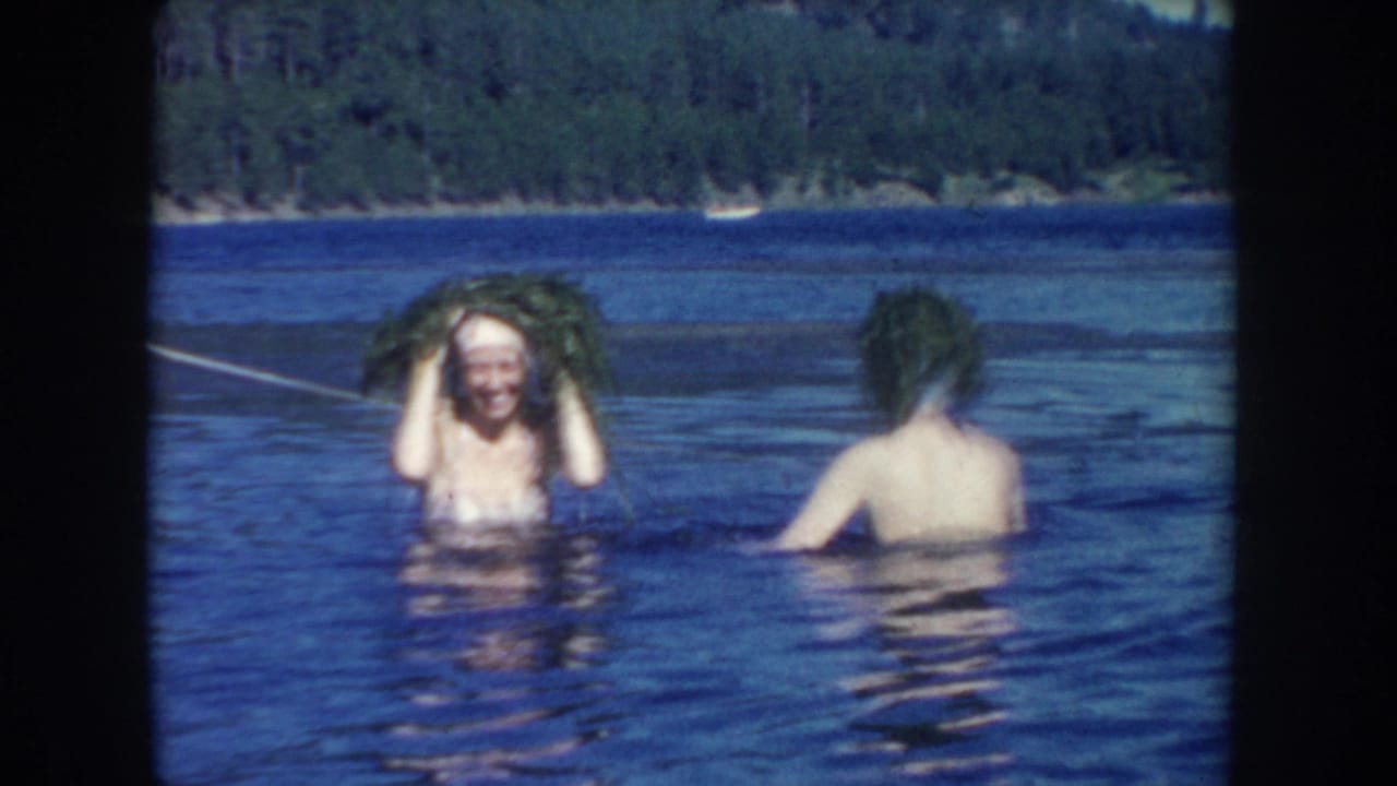 1954 South Dakota home-movie frame showing two women waist-deep in a lake, one smiling toward the camera while holding wet hair.