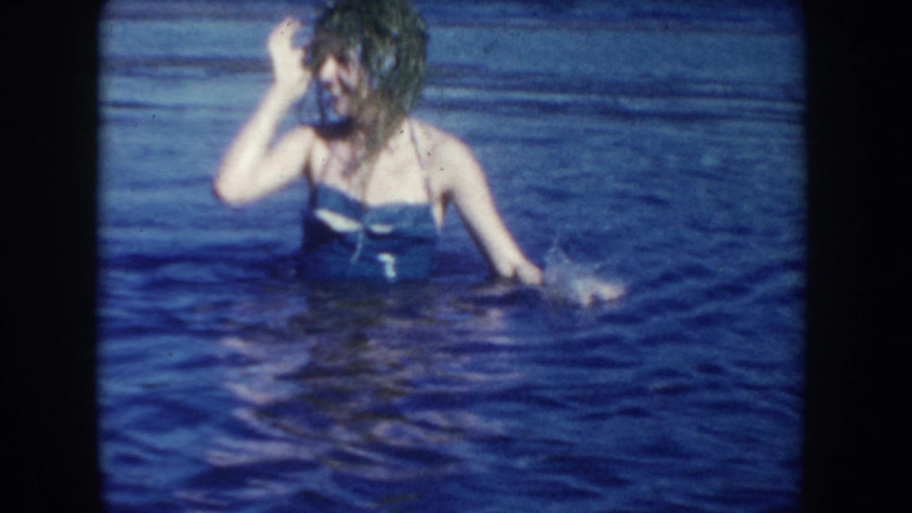 1954 South Dakota frame of a woman in a blue swimsuit standing in lake water and touching her wet hair, with open water behind her.