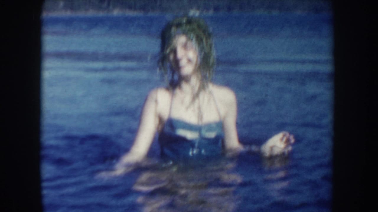 1954 South Dakota home-movie still of a woman in a blue swimsuit chest-deep in lake water, with soft focus and saturated blue tones.