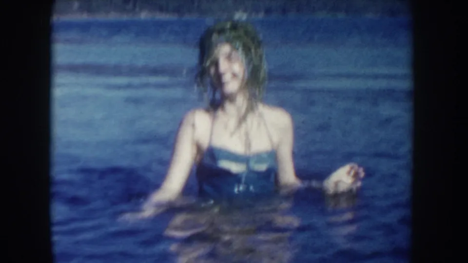 1954 South Dakota home-movie still of a woman in a blue swimsuit chest-deep in lake water, with soft focus and saturated blue tones.