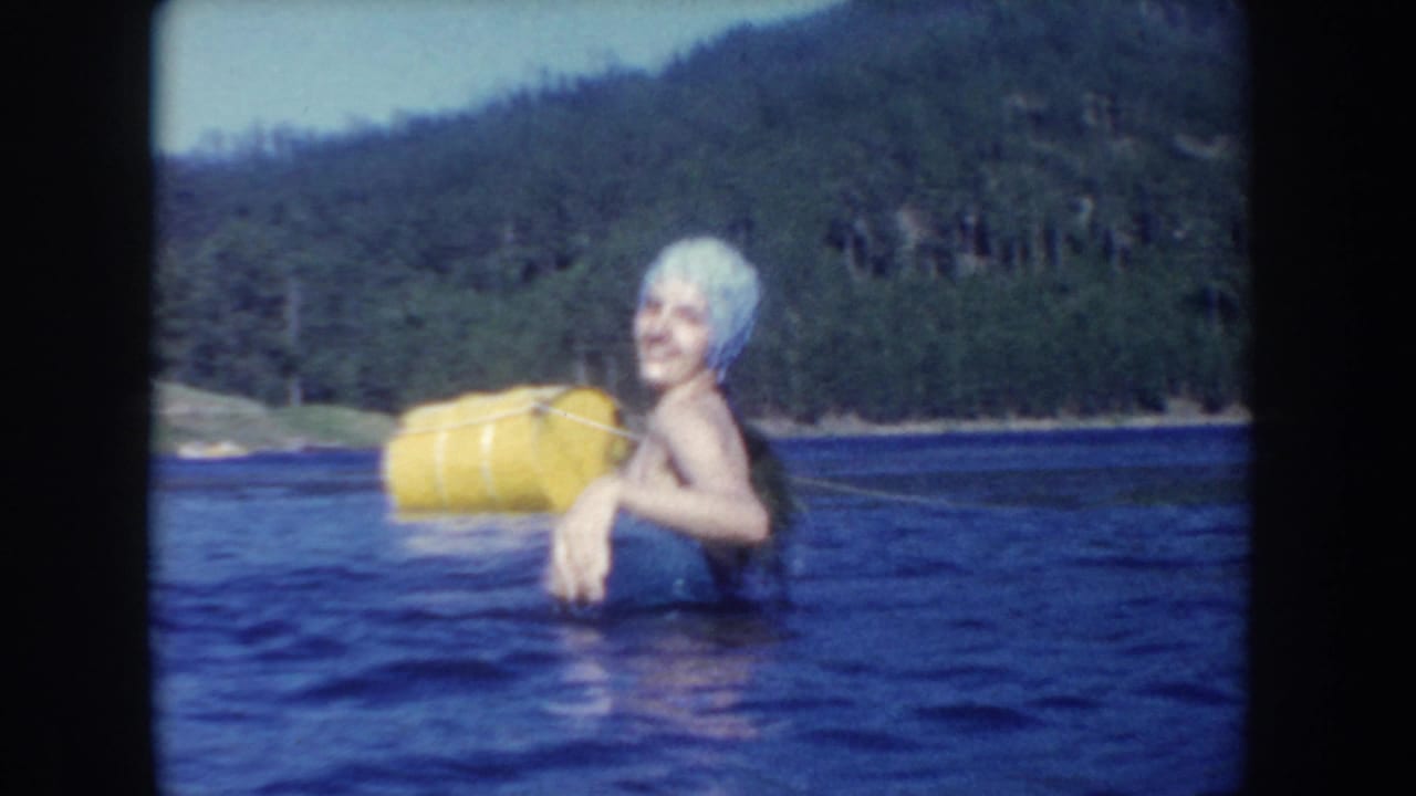 1954 South Dakota frame showing a smiling woman in a pale swim cap beside a yellow flotation object, with wooded hills across the lake.
