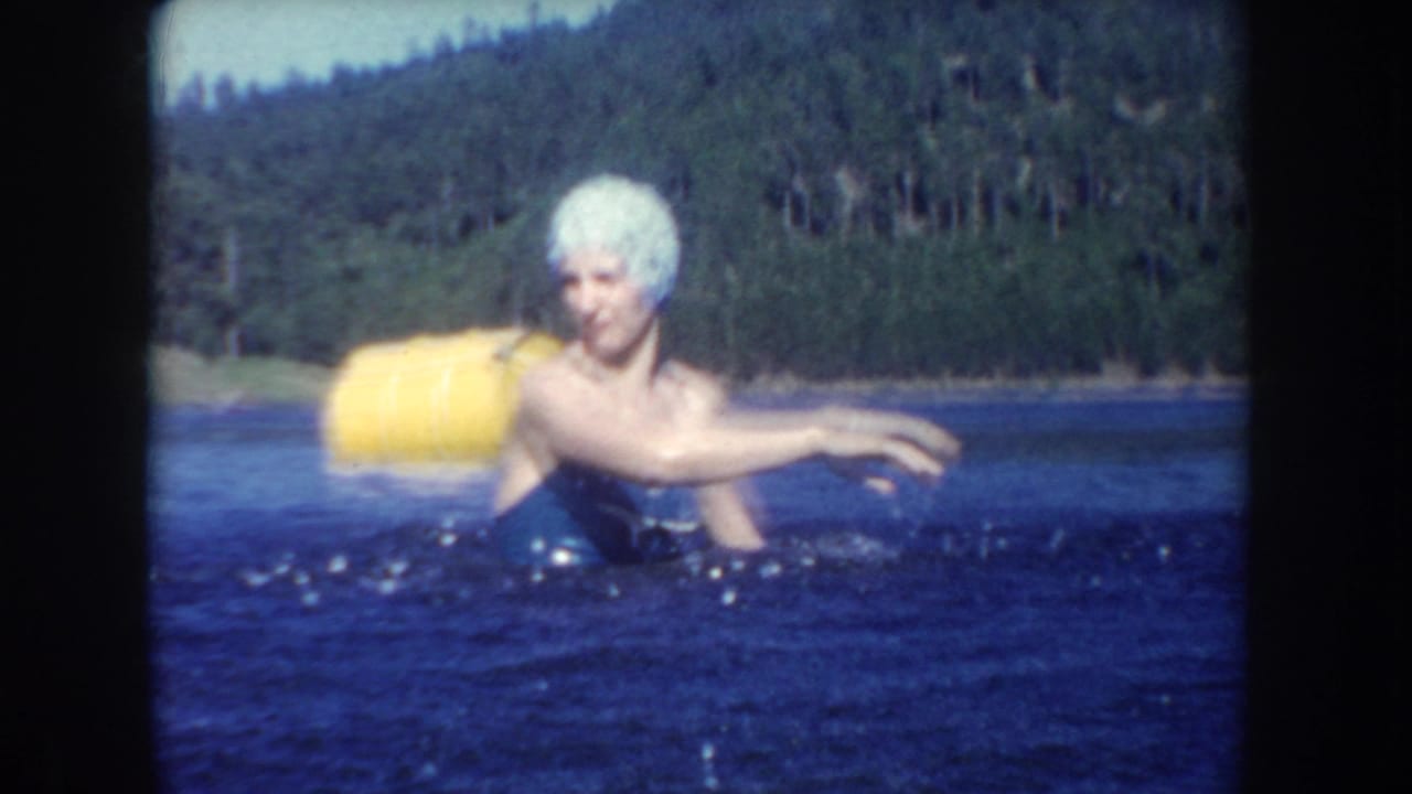 Woman in a white swim cap swims near a yellow buoy with forested shore behind her.