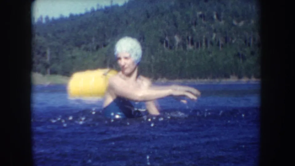 1954 South Dakota home-movie frame of a woman in a white swim cap and dark swimsuit swimming beside a yellow barrel buoy.