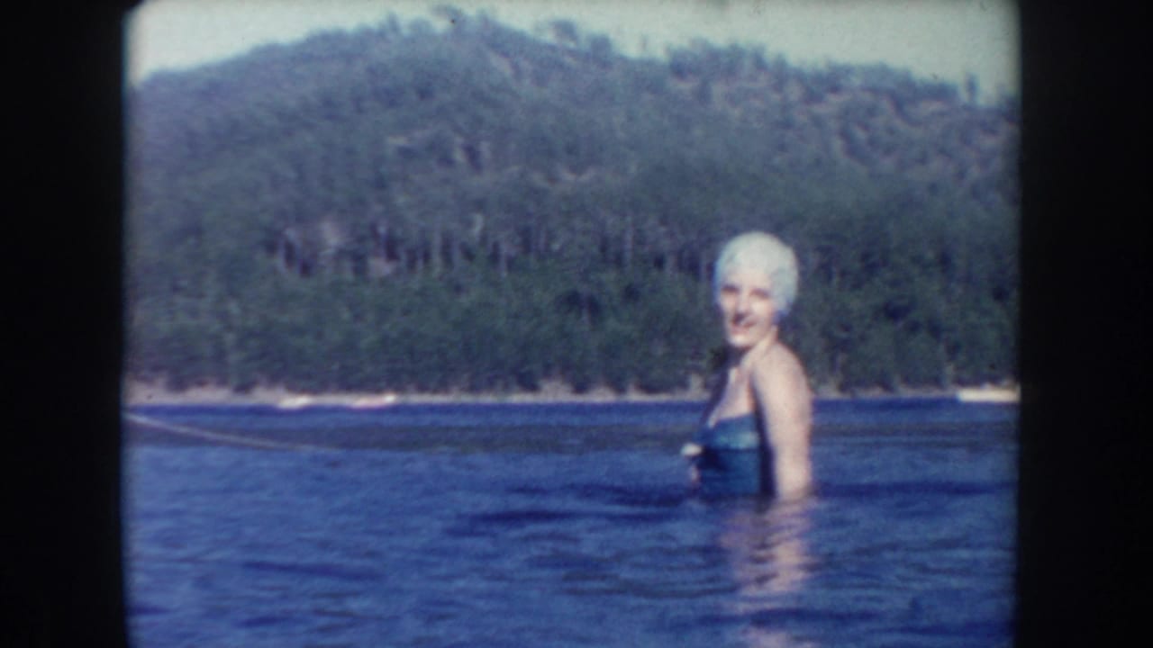 1954 South Dakota 8mm frame showing a woman in a white swim cap standing in lake water before a forested shoreline.