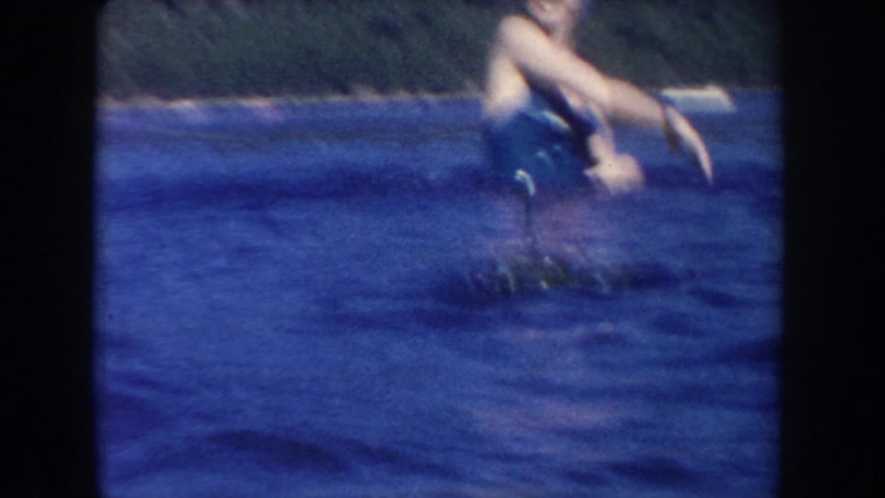 Soft 1954 South Dakota lake frame with a partially cropped swimmer reaching over the water near floating vegetation.