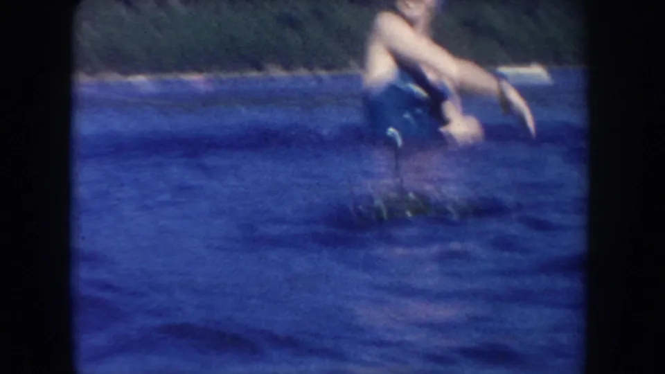 Soft 1954 South Dakota lake frame with a partially cropped swimmer reaching over the water near floating vegetation.