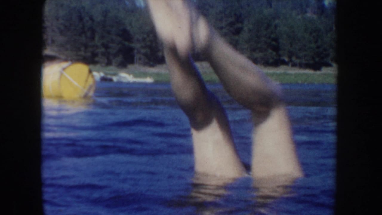 1954 South Dakota lake frame showing legs splashing upward from the water, with a yellow buoy and forested shoreline in the background.