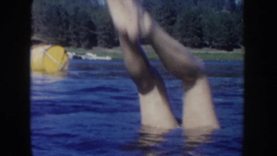 1954 South Dakota lake frame showing legs splashing upward from the water, with a yellow buoy and forested shoreline in the background.