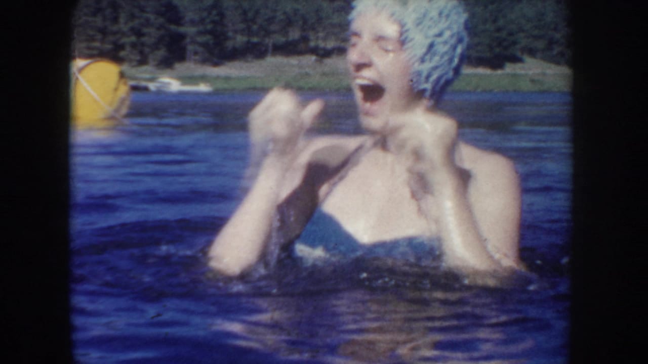 1954 South Dakota 8mm frame showing a laughing swimmer in lake water, with a yellow buoy and tree-lined shoreline behind them.