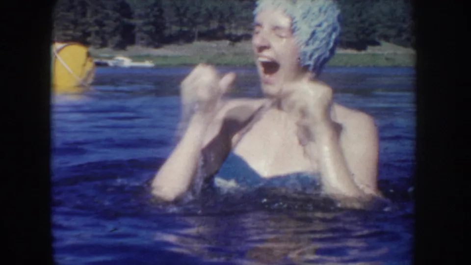 1954 South Dakota 8mm frame showing a laughing swimmer in lake water, with a yellow buoy and tree-lined shoreline behind them.