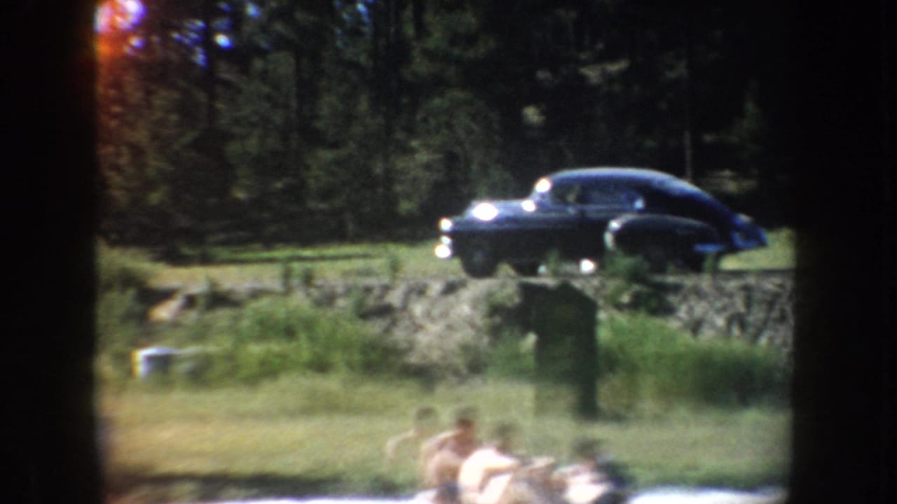 A dark 1950s-style sedan sits on a grassy rise above a lakeside swimming area in South Dakota home-movie footage.
