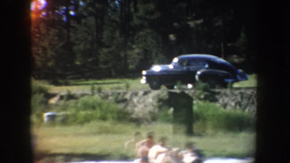 A dark 1950s-style sedan sits on a grassy rise above a lakeside swimming area in South Dakota home-movie footage.