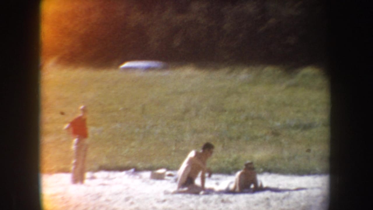 People relax on a sandy lakeshore below a grassy hillside, with soft focus and orange film flare from the 8mm scan.
