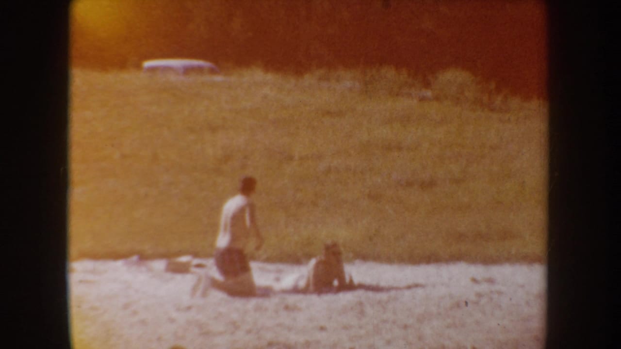 Two beachgoers remain near the lakeshore as the frame shifts warm orange, showing the home-movie film character clearly.