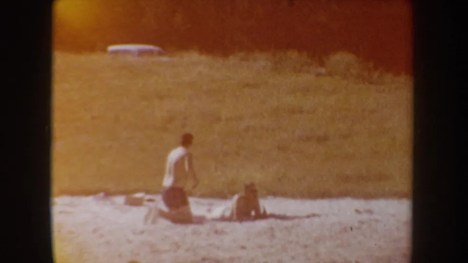 Two beachgoers remain near the lakeshore as the frame shifts warm orange, showing the home-movie film character clearly.
