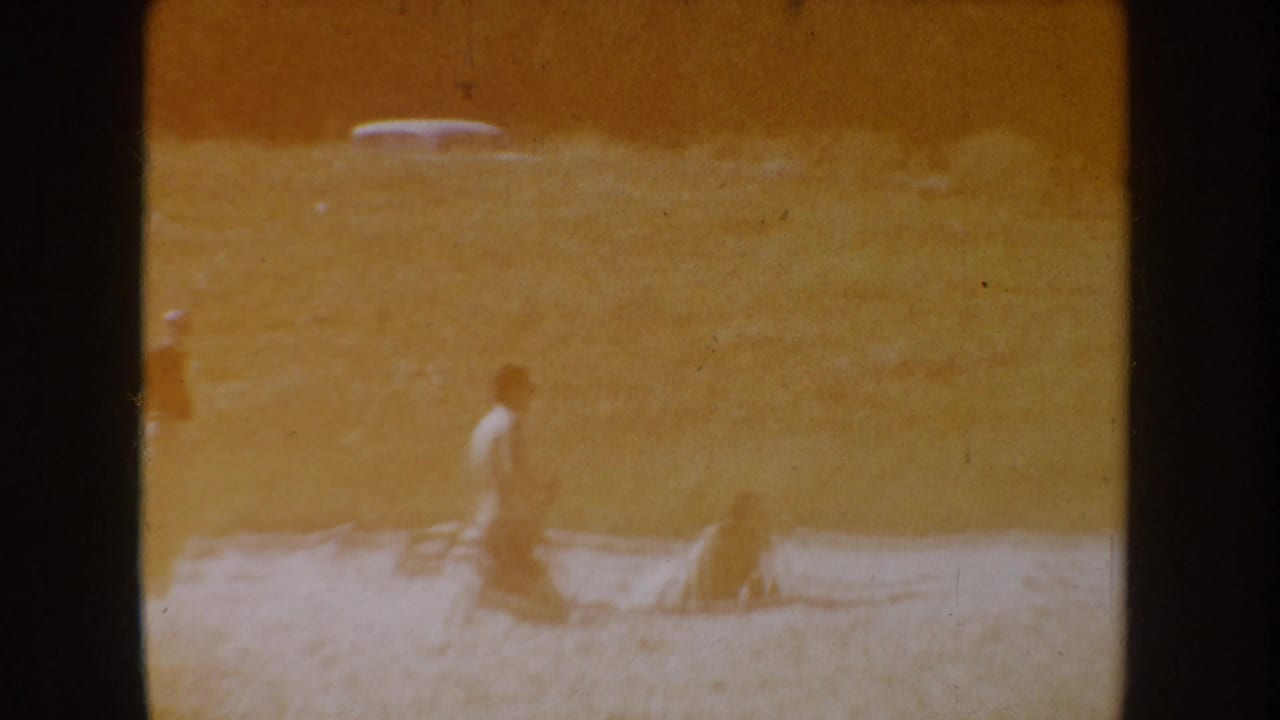 South Dakota 1954 home-movie frame showing two distant people near a lakeside shore, one standing or walking and one lower to the ground.