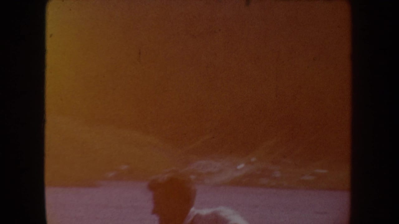 Yellow-tinted 8mm frame with a faint person-like silhouette in the lower foreground and indistinct outdoor ground beyond.