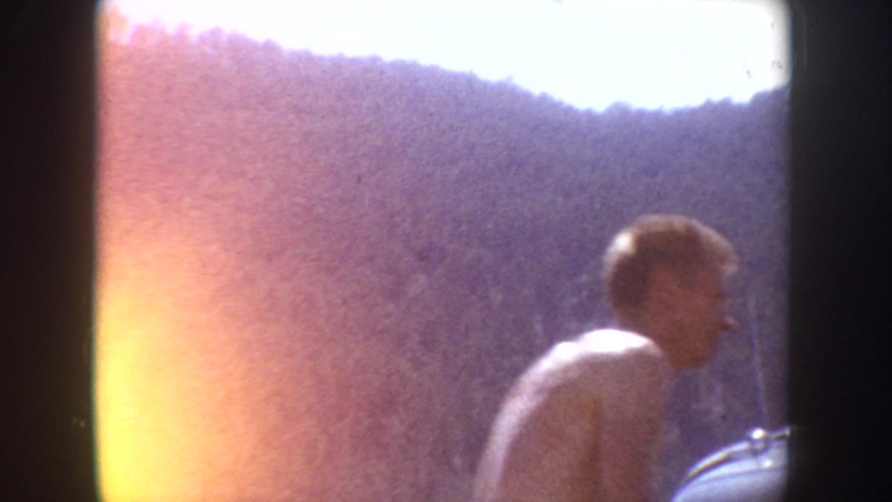 1954 South Dakota home-movie frame showing a young man near a parked car, with wooded hills and strong film flare at the edge.