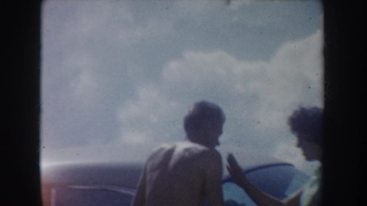 1954 South Dakota lakeside home-movie frame with two friends near a car, partly silhouetted against sky and hills.