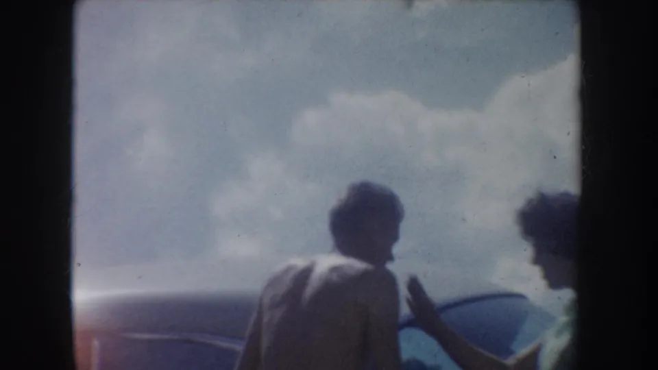 1954 South Dakota lakeside home-movie frame with two friends near a car, partly silhouetted against sky and hills.