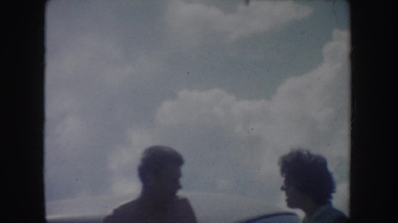 Soft 8mm frame from the South Dakota lake outing, showing two friends in shadow beside a car with clouds behind them.