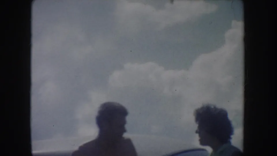 Soft 8mm frame from the South Dakota lake outing, showing two friends in shadow beside a car with clouds behind them.