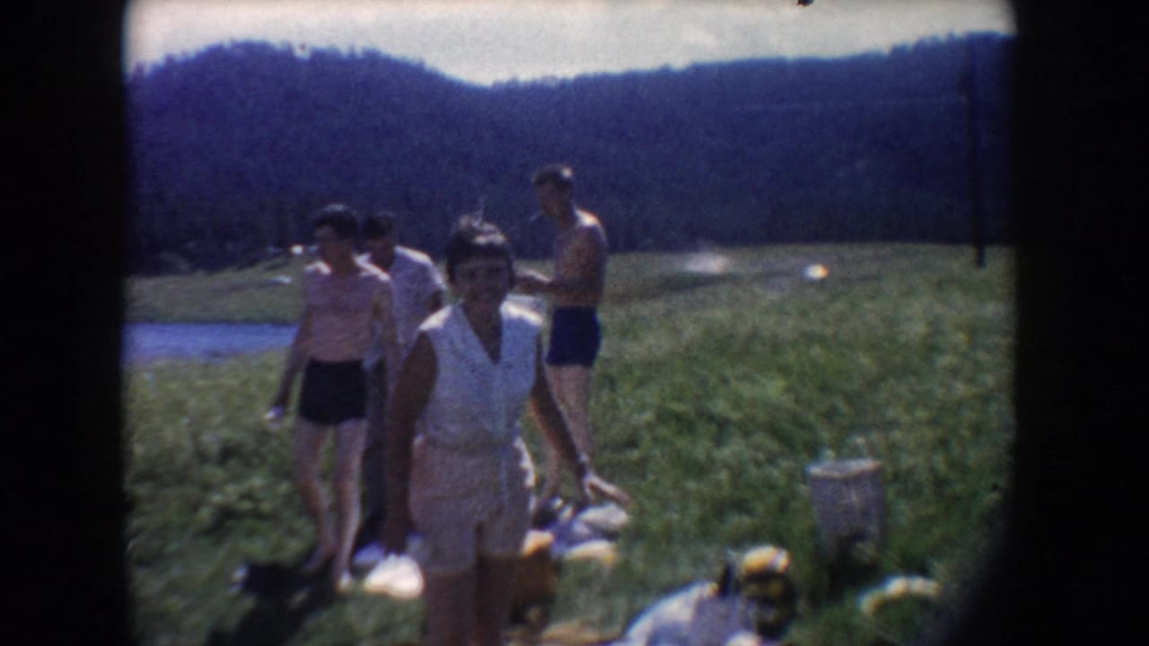 1954 South Dakota home-movie frame showing friends in swimwear and summer clothes crossing a grassy lakeside meadow.
