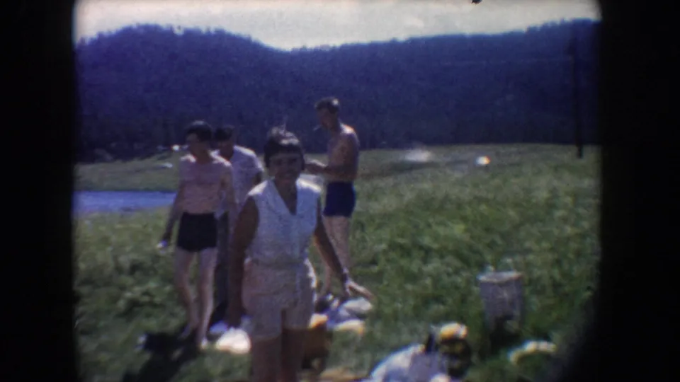 1954 South Dakota home-movie frame showing friends in swimwear and summer clothes crossing a grassy lakeside meadow.