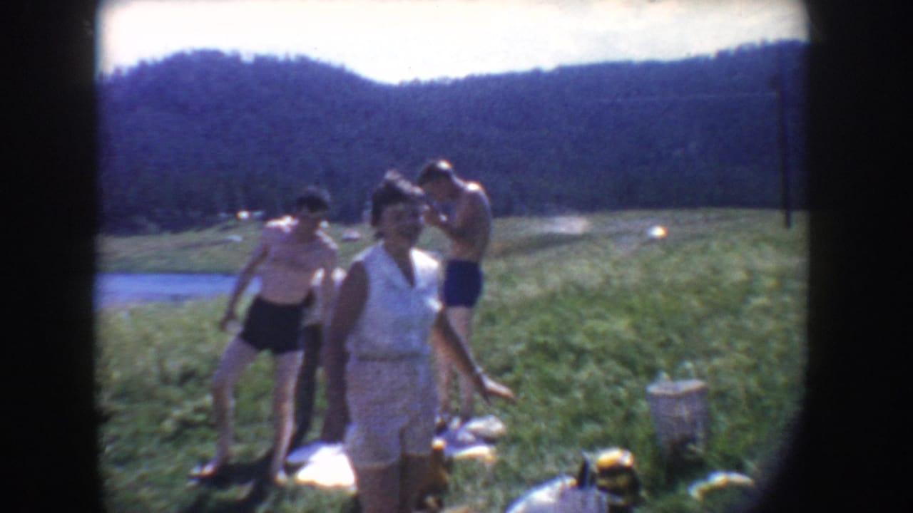 1954 South Dakota home-movie frame of adults in summer clothing near picnic blankets on grass beside a lake and wooded hills.