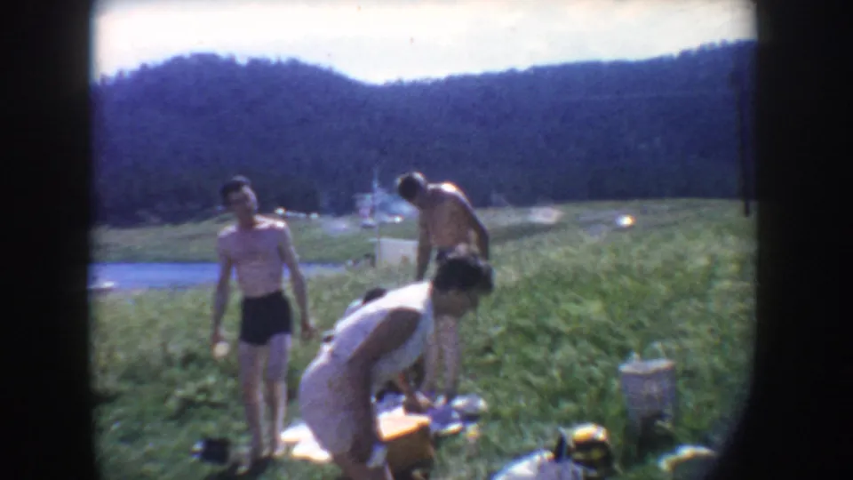 Adults on a South Dakota lakeshore in 1954 appear to pack or arrange picnic items near blankets, water, and wooded hills.