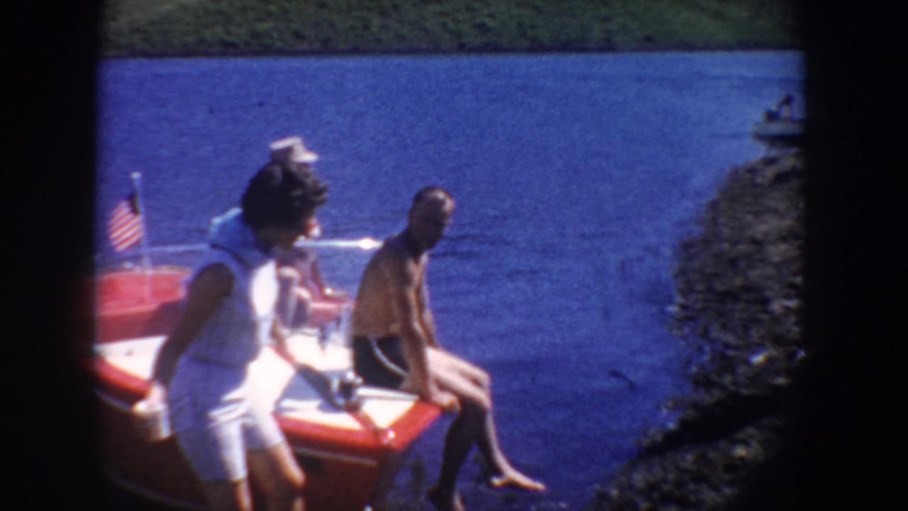 1954 South Dakota lake scene with adults near a red-and-white motorboat, blue water, rocky shore, and a small American flag.