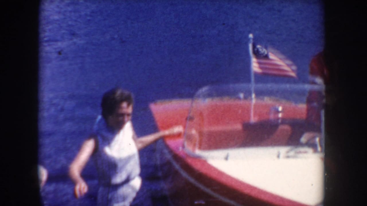 A woman in a sleeveless white outfit stands beside a red-and-white motorboat flying an American flag on a South Dakota lake.