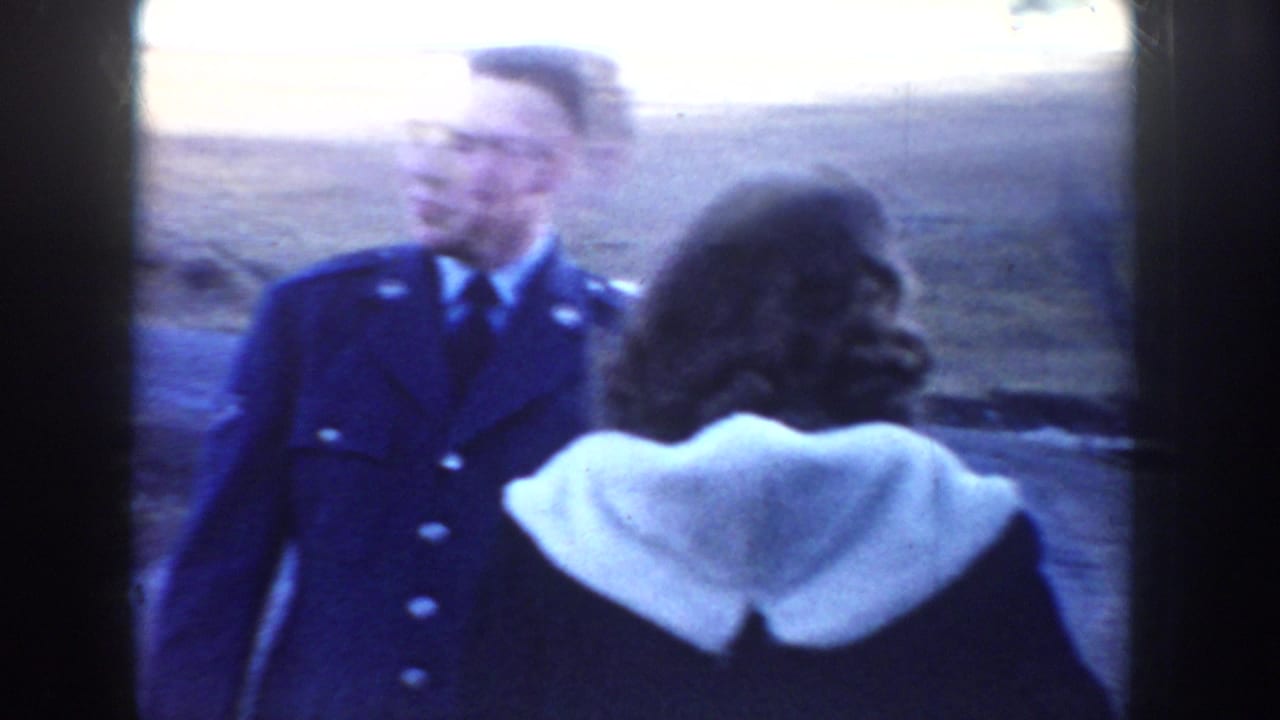 In 1955 Idaho home-movie footage, a man in a blue military-style uniform stands near a woman in a dark coat along a rural road.