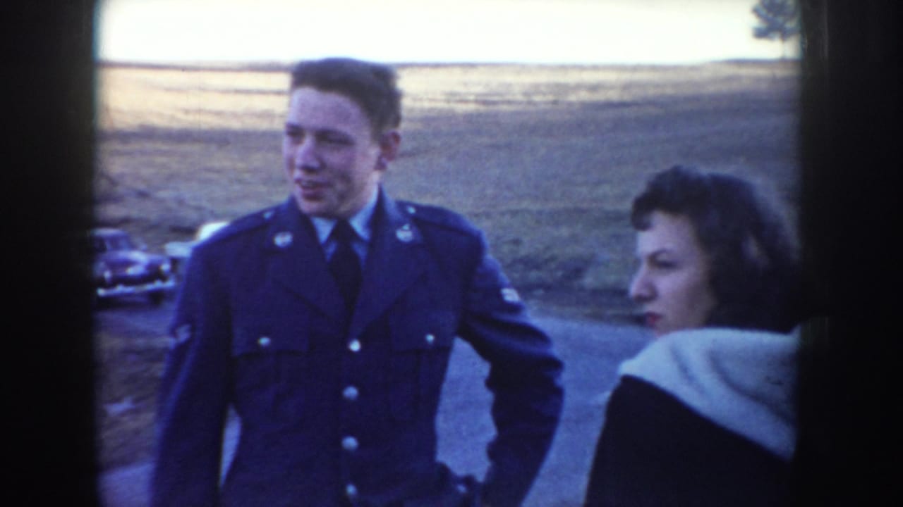 The uniformed man and woman appear to speak or look toward each other beside a rural Idaho road, with an older car in the background.