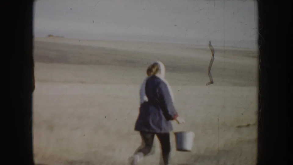 A person in a dark coat walks away across the dry Idaho field carrying a light bucket, with the horizon in the distance.