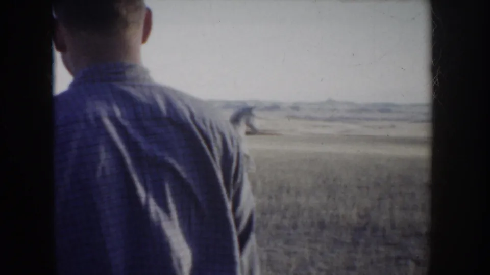 A man in a light shirt is seen from behind in the foreground, with horses and the Idaho landscape visible beyond him.