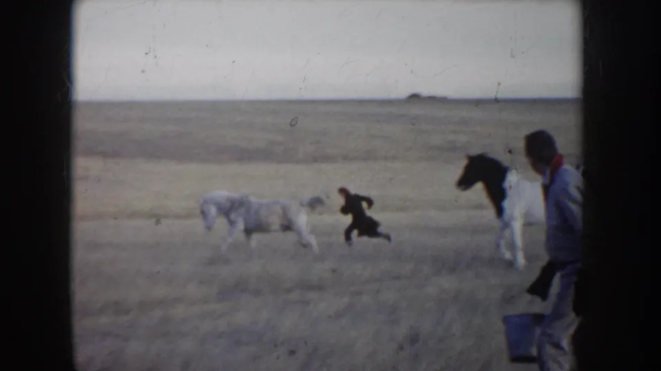 1955 Idaho 8mm frame showing a person running near horses on open prairie, with another adult partly visible at the edge.