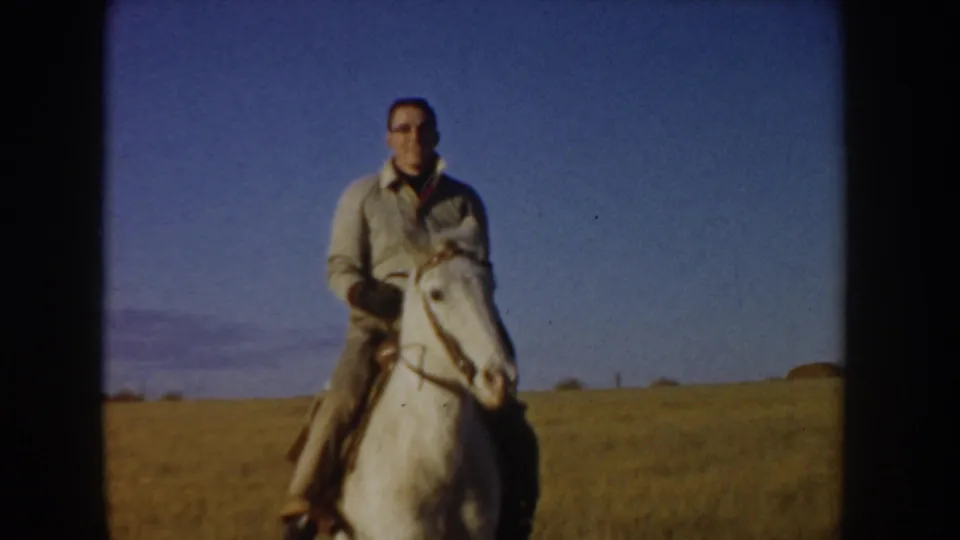 Adult man rides a white horse across Idaho prairie in a 1955 8mm home-movie frame, with blue sky and distant hills behind him.