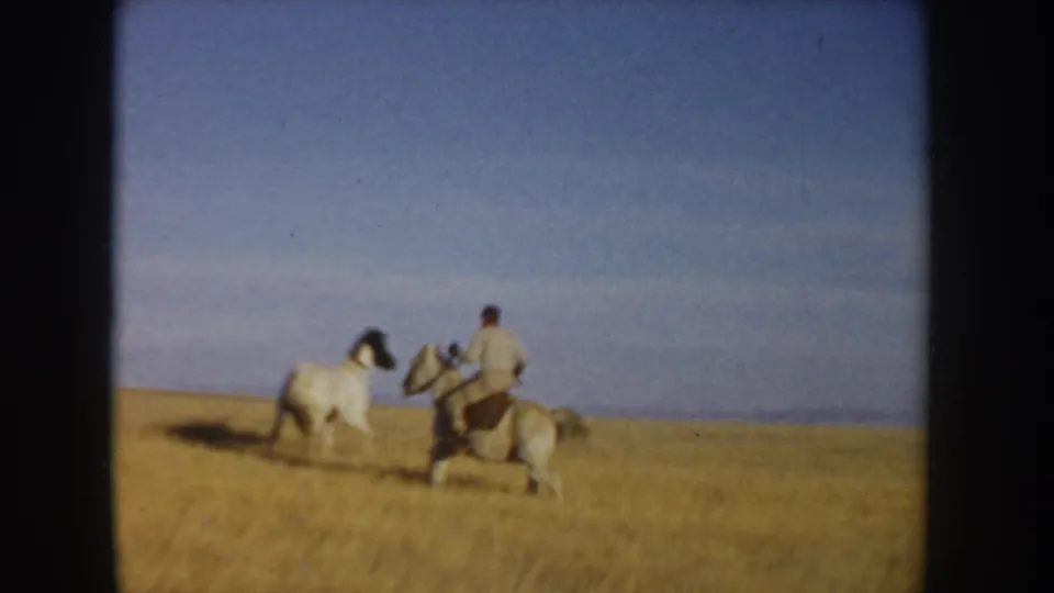 Two riders on horses cross open Idaho grassland in a 1955 8mm frame, with a wide blue sky and low horizon.