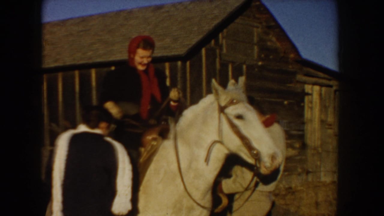 Woman in a red headscarf seated on a white horse beside a wooden barn.